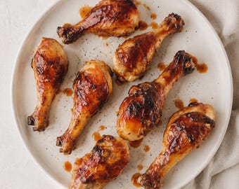 May include: A plate of seven roasted chicken drumsticks. The chicken has a glossy, caramelized exterior with a rich brown color. The plate is white, and the background is a neutral color. A few drops of sauce are visible on the plate.