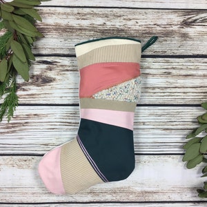 May include: A patchwork Christmas stocking in shades of pink, beige, and green. The toe and heel are pink, with a dark green section and a striped beige section. The stocking has a green loop at the top. The design is a mix of textures and patterns.