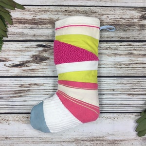 Può includere: Una calza di Natale fatta a mano con un design patchwork. La calza presenta strisce di tessuti rosa, bianchi, gialli e blu. La parte superiore ha un bordo bianco e un anello blu per appenderla. Sfondo in legno rustico.