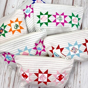May include: A collection of rectangular zipped pouches made from off-white fabric with thin horizontal grey stripes. Each pouch features a row of colourful star-shaped appliques, with small patterned fabric squares centred within each star point.