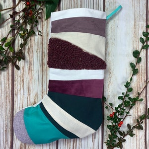 May include: A handmade Christmas stocking with a patchwork design. The stocking features various fabrics in shades of white, purple, teal, and burgundy. A textured burgundy fabric is sewn diagonally across the center. A teal loop is attached at the top.