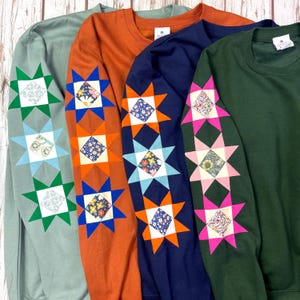 May include: Four long-sleeve crewneck sweatshirts in sage green, burnt orange, navy blue, and forest green. Each sleeve features a vertical row of three star-shaped patches with floral patterned centres in contrasting colours and geometric points.