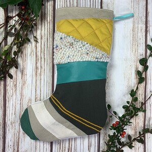 May include: A handmade Christmas stocking with a patchwork design. The stocking features sections of mustard yellow quilted fabric, teal leather, and a textured white and speckled material. The toe is striped with green, white, and gray. A teal loop is attached for hanging.