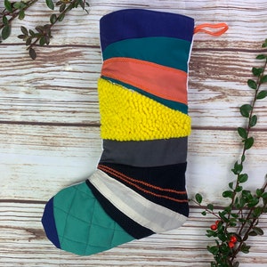 Puede incluir: Un calcetín navideño hecho a mano con un diseño de patchwork. El calcetín presenta varias telas en tonos de azul, verde, naranja, amarillo, gris y negro. Tiene una puntera acolchada y un lazo naranja para colgar. El diseño es una mezcla de texturas y patrones.