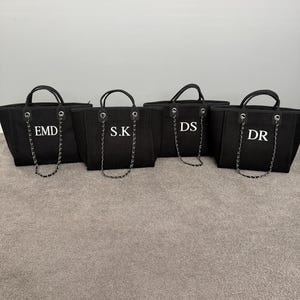 May include: Four black tote bags with two top handles and a decorative silver-toned chain strap. Each bag features white embroidered initials on the front, reading EMD, S.K, DS, and DR respectively. The bags have a structured rectangular shape.