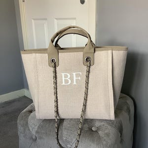 Könnte beinhalten: Beige Tragetasche mit Kettenriemen und Tragegriffen. Die Tasche hat die weißen Initialen "BF" auf der Vorderseite. Die Tasche steht auf einem gepolsterten, grauen Hocker.
