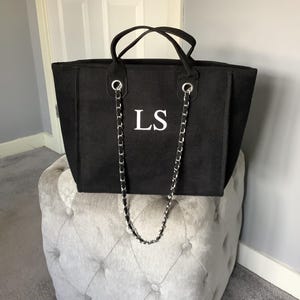 May include: A black tote bag with a textured fabric finish, featuring the white letters LS centred on the front. The bag includes two black top handles and a long silver chain shoulder strap that hangs down the front, attached via silver grommets.
