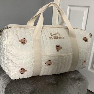 May include: A white quilted duffel bag featuring embroidered brown teddy bear faces scattered across the surface. The bag includes beige fabric handles and the text Baby Williams embroidered in brown thread on the front center panel.