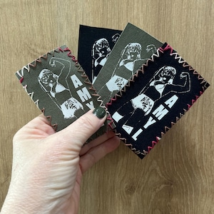May include: Three iron-on patches with a white silhouette of a woman flexing her bicep and the word "AMY" written vertically. The patches are on a wood background. The patches are black, dark green, and light green.