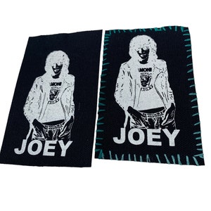 May include: Two black fabric patches with white stitching around the edges. Each patch features a white silhouette of a person wearing a leather jacket and a t-shirt with the text "L'MONE" and "ROYCE" printed on it. The text "JOEY" is printed in white at the bottom of each patch.