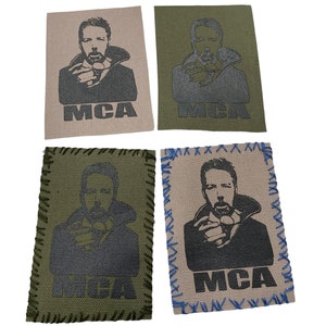 May include: Four fabric patches with a black and white image of a man with a beard and a black shirt. The patches are in two different colors: beige and green. The patches have the text "MCA" printed on them.
