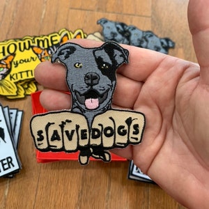 May include: A gray and black pit bull dog with a pink tongue is shown with its paws in a fist shape. The paws are embroidered with the text "SAVEDOGS".