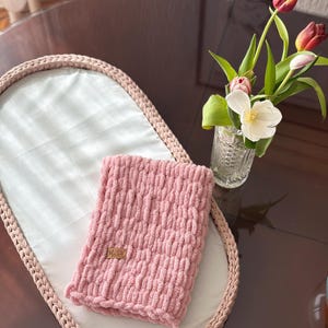 May include: A pink, chunky-knit blanket with a small wooden tag, on a white fabric-lined, oval tray with a braided edge. A vase of tulips sits nearby on a dark, glossy surface.