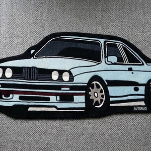 May include: A car-shaped rug in light blue and black, resembling a classic coupe. The rug features detailed design elements like headlights, wheels, and a black outline. The rug is placed on a gray surface.