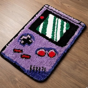 Könnte beinhalten: Ein lila, schwarzer und roter Teppich in Form einer klassischen Handheld-Spielkonsole. Der Teppich hat einen Bildschirm mit grünen und weißen Streifen, ein Steuerkreuz, Knöpfe und einen Power-Knopf. Der Teppich ist etwa 76 cm lang und 51 cm breit.