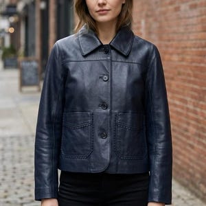 May include: A dark navy blue leather jacket featuring a classic collar, a front button closure with four visible buttons, and two front patch pockets with decorative scalloped trim along the edges. The jacket has long sleeves and a straight hem.