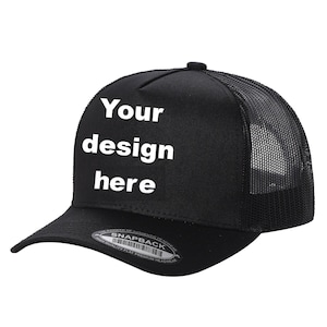 May include: Black trucker hat with a mesh back and a curved brim. The front panel displays the text "Your design here" in white, offering a customizable design space. The hat features a snapback closure for adjustable sizing.