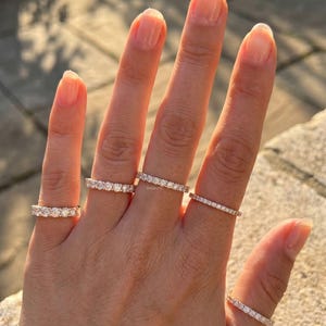 May include: A hand displays five rose gold rings, each adorned with sparkling gemstones. The rings vary in design, showcasing different gemstone arrangements and band widths. The sunlight highlights the details of the jewelry.