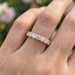 May include: A gold ring with a band set with a row of round-cut diamonds. The ring is shown against a backdrop of blurred green leaves and pink flowers, emphasising the ring's sparkle and design.