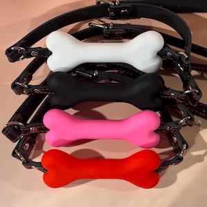 Puede incluir: Cuatro collares para perros con juguetes masticables en forma de hueso en blanco, negro, rosa y rojo. Los collares son de cuero negro con hebillas plateadas y costuras rojas.