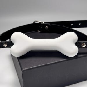 May include: A black leather collar with a white bone-shaped centerpiece. The collar has a silver buckle and rivets. The bone is smooth and solid, contrasting with the textured leather. The item is presented on a black box.