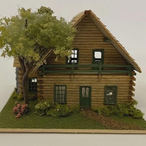 May include: A miniature wooden cabin with a green trim, a brown roof, and a small tree. The cabin has multiple windows and a green door. The model sits on a green base with a path leading to the door.