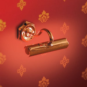 May include: A gold-colored picture light fixture with a curved arm and a rectangular light housing. The fixture is mounted on a red wall with a repeating gold floral pattern. The light is designed to illuminate artwork.