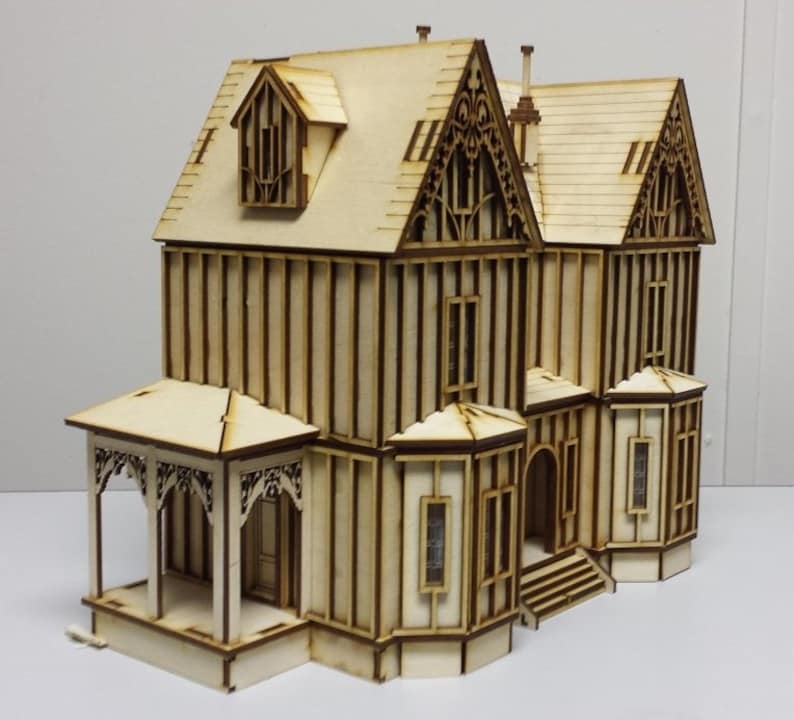 May include: A wooden model of a two-story house with a porch, intricate details, and a gabled roof. The model is made of light brown wood and has a white background.