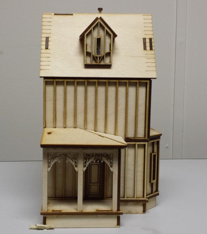 May include: A wooden model of a two-story house with a porch and a dormer window. The model is made of light brown wood and has a simple design.