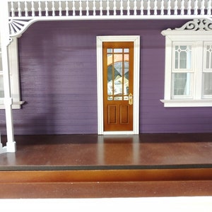 May include: A dollhouse porch with a purple exterior, white trim, and a brown wooden door with a stained glass window. The porch has a brown wooden floor and a white railing.