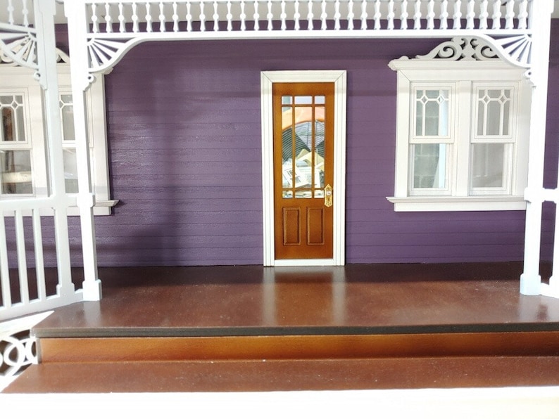 May include: A dollhouse porch with a purple exterior, white trim, and a brown wooden door with a stained glass window. The porch has a brown wooden floor and a white railing.
