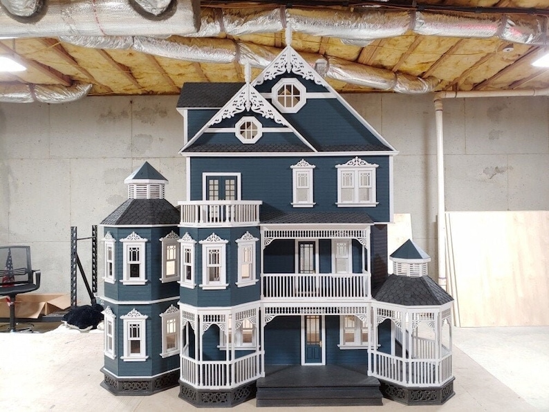 May include: A detailed dollhouse model with a blue exterior, white trim, and a black roof. The dollhouse has a wraparound porch with white railings and a front door with a glass window. The dollhouse has a second-story balcony with white railings and a window with a white frame.
