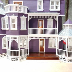 May include: A purple dollhouse with white trim and a white wraparound porch. The dollhouse has a gabled roof and a cupola on top. The porch has white railings and a white picket fence.