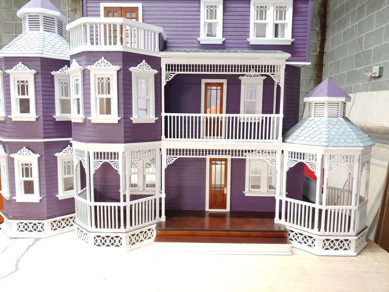 May include: A purple dollhouse with white trim and a white wraparound porch. The dollhouse has a gabled roof and a cupola on top. The porch has white railings and a white picket fence.