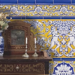 Puede incluir: Escena decorativa con un mueble de madera con espejo, candelabros y un jarrón azul y blanco con flores. El fondo muestra azulejos estampados amarillos, azules y blancos con diseños de querubines y florales, creando una estética vintage.