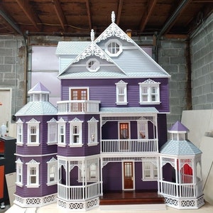 May include: A purple dollhouse with white trim and a gray roof. The dollhouse has a wraparound porch with white railings and a balcony on the second floor. The dollhouse has several windows and doors, and a small cupola on the roof.