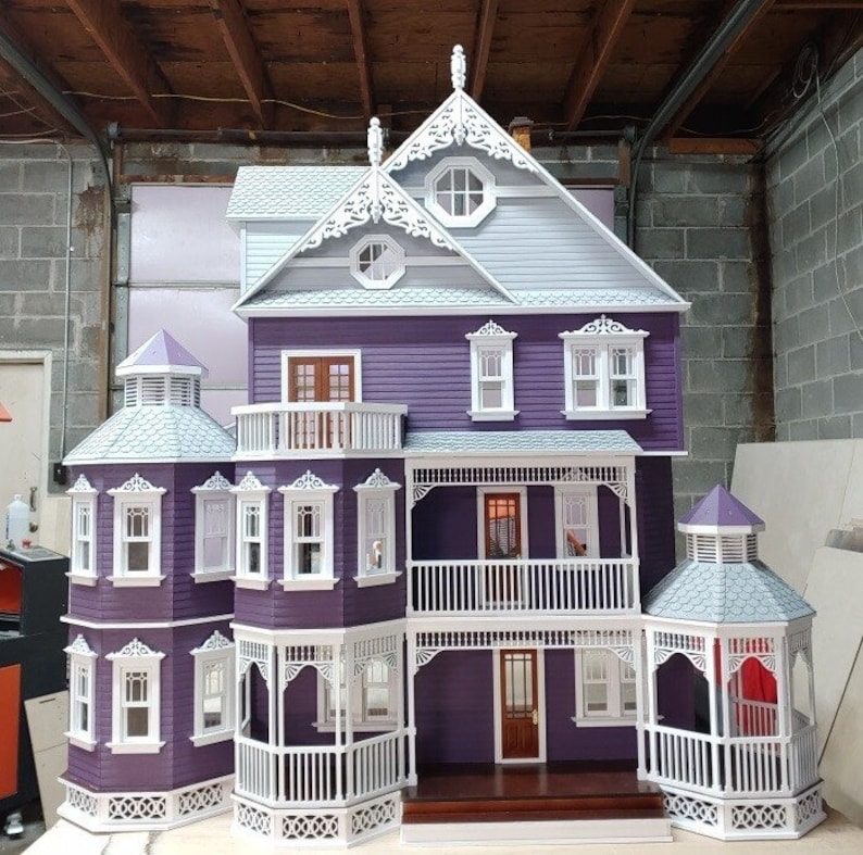 May include: A purple dollhouse with white trim and a gray roof. The dollhouse has a wraparound porch with white railings and a balcony on the second floor. The dollhouse has several windows and doors, and a small cupola on the roof.