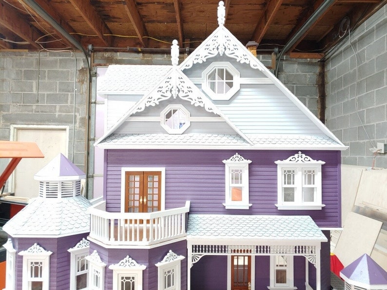May include: A purple and white dollhouse with a detailed exterior, including a balcony, porch, and decorative trim. The roof is white with a gray shingle pattern.