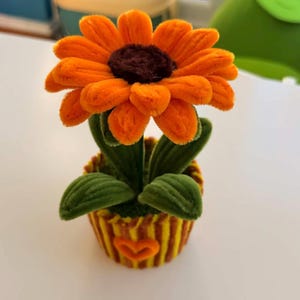 Può includere: Un girasole arancione fatto a mano con un centro marrone, foglie verdi e un vaso a righe gialle e arancioni. Il fiore è fatto di un materiale morbido e soffice e il vaso ha un design a forma di cuore.