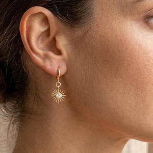 May include: A gold hoop earring featuring a sunburst charm with a small, clear, round-cut stone at its center. The charm hangs from a thin, circular gold hoop that is secured through the earlobe, showcasing a radiant, star-like design.