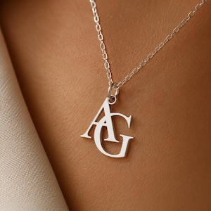 May include: A silver-toned necklace featuring a pendant with the interlocking capital letters A and G. The pendant is suspended from a delicate, thin silver-toned chain with small, uniform rectangular links that connect to a circular jump ring.