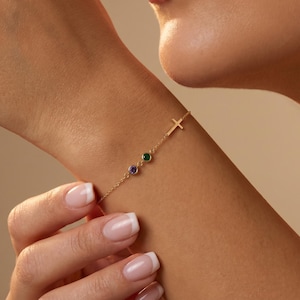 May include: Gold bracelet featuring a cross charm and two gemstones, one green and one purple. The delicate chain bracelet is worn on a wrist, showcasing the jewelry's design and color against the skin.