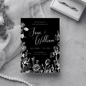 Op de afbeelding: Een zwarte trouwkaart met witte bloemenillustraties en tekst. De uitnodiging luidt "Please join us for the wedding of Jane & William" met datum en tijd. Een witte doos met twee ringen staat op de achtergrond.