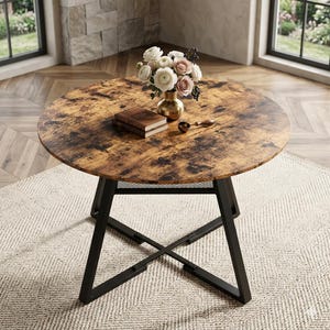 Peut inclure: Table ronde en bois avec une finition marron vieilli, soutenue par des pieds en métal noir. Un vase de fleurs, un livre et une cuillère sont sur la table. La table est posée sur un tapis beige.