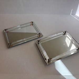 May include: Two rectangular mirrored trays featuring clear glass rod borders and metallic gold-toned corner accents. Each tray has a reflective surface framed by parallel glass rods that create a layered effect, providing a clean and polished look.