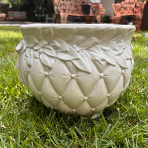 May include: A cream-colored, decorative planter with a quilted pattern and leaf-like detailing around the rim. The planter sits on short feet and is designed for outdoor use. The planter is sitting on green grass.