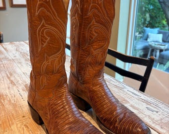 May include: A pair of tan leather cowboy boots featuring a textured lizard-skin pattern on the foot and a smooth leather shaft with intricate white stitching details. The boots have a pointed toe, a tall shaft, and a stacked wooden heel.