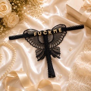 Peut inclure: Un string en dentelle noire présentant un motif en forme de papillon avec des bords festonnés et une fine ceinture élastique. Le mot FUCK ME est écrit en lettres dorées incrustées de strass sur le haut du motif papillon en dentelle.
