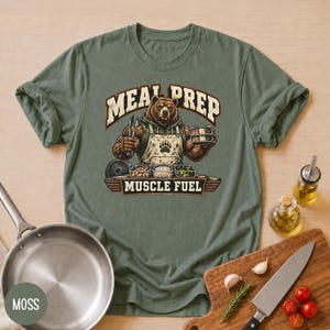 Könnte beinhalten: Ein moosgrünes T-Shirt mit einer Grafik eines muskulösen braunen Bären in einer Schürze, der eine Zange und einen Essensbehälter hält. Der Text MEAL PREP steht über dem Bären und MUSCLE FUEL darunter, umgeben von drei weiteren Essensbehältern.