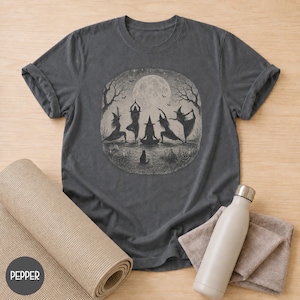 Puede incluir: Una camiseta gris carbón con un gráfico circular de cinco brujas con sombreros puntiagudos realizando posturas de yoga bajo una luna llena. La ilustración incluye árboles desnudos, un pequeño gato negro y calabazas. Mangas cortas y cuello redondo.